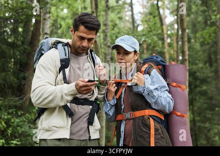 Young adult Caucasian man and young adult Caucasian woman standing in forest using GPS device while hiking, both carrying backpacks and outdoor gear, surrounded by trees and greenery Stock Photo