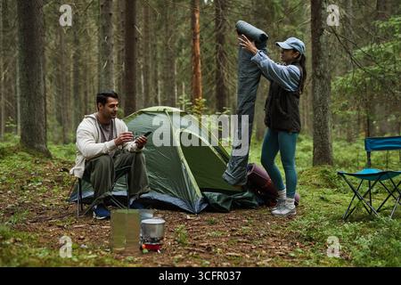 Caucasian young adult man sitting near tent using smartphone while Caucasian young adult woman standing nearby holding rolled sleeping bag in forest campsite setting Stock Photo