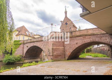 Large stone bridge with a church in the background under a cloudy sky, decorated with colourful flags, small town of Perle Calw, Black Forest, Germany Stock Photo
