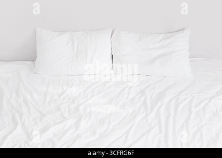 White bed with white linens with two pillows close up against the background of a white wall Stock Photo
