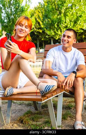 Young couple relaxing on bench, woman using smartphone, man smiling Stock Photo
