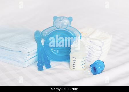 Baby tableware set. milk bottle, feeding nibbler, blue cutlery, spoon, fork, diapers, disposable diapers on the white bed background. front view Stock Photo