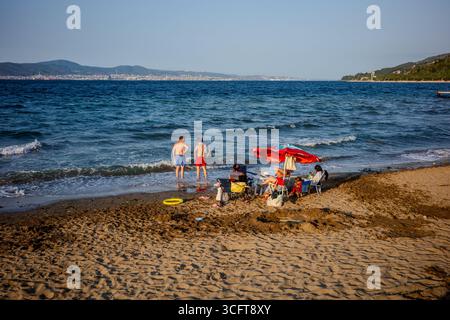 Beachgoers sit under an umbrella and enjoy the shoreline while two men stand in the water along the coast of Karamürsel, Kocaeli, Türkiye. The Marmara Stock Photo