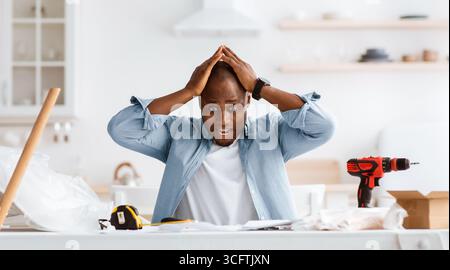 Frightened handyman having problems in assembling new furniture, sitting in kitchen interior Stock Photo