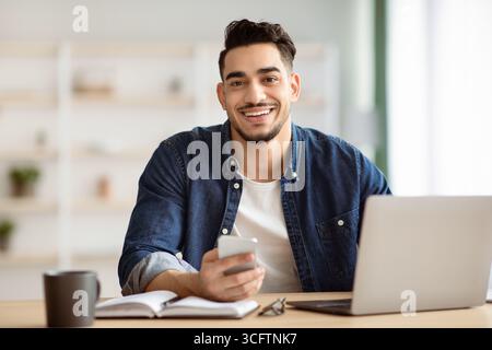 Smiling arab guy using smartphone while sitting at workdesk Stock Photo