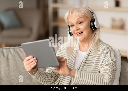 Smiling old lady in headset using tablet sitting on sofa Stock Photo