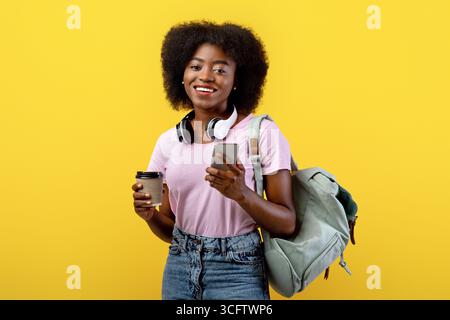 Positive african american woman with backpack and smartphone holding coffee to go over yellow background Stock Photo