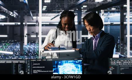 Team of data center engineers looking over paperwork and using deep learning algorithms to analyze datasets. Employees in multiethnic server farm using AI to recognize data patterns, camera A Stock Photo