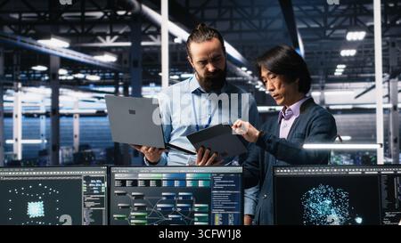 Data center manager supervising developer using device to monitor neural network AI LLMs. Team leader checking progress done by worker overseeing servers powering machine learning processes, camera A Stock Photo