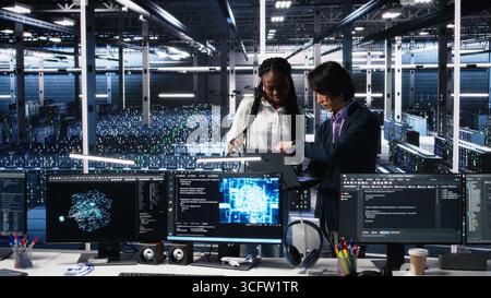 Team of data center engineers looking over paperwork and using deep learning algorithms to analyze datasets. Employees in multiethnic server farm using AI to recognize data patterns, camera A Stock Photo