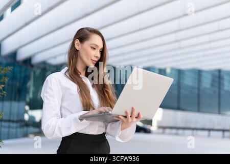 Confident female manager using laptop in front of office building. Young businesswoman wearing white shirt. Woman is holding netbook outside workplace Stock Photo