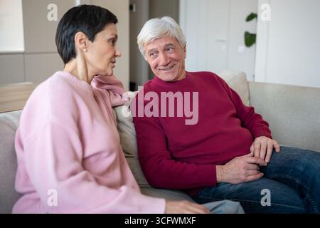 Joyful retired couple home rest. Elderly husband telling stories to attentive wife on sofa at home Stock Photo
