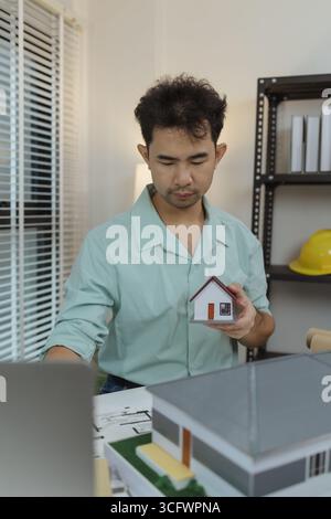 Asian male architect holding a miniature house model while working at his desk. Surrounded by blueprints and architectural tools, he appears to be pla Stock Photo