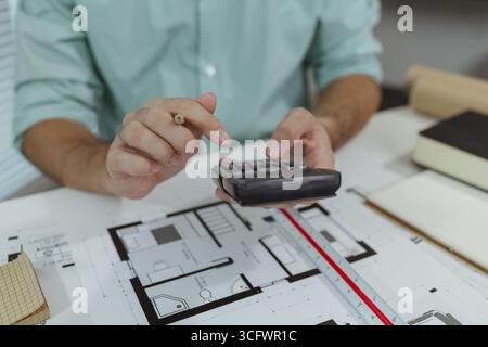 An architect in a green shirt is sitting at a white desk, using a calculator while reviewing printed house floor plans. The image highlights the works Stock Photo