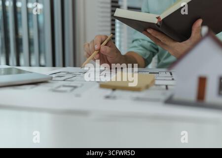 Architect reviewing house floor plan while holding a notebook and pencil. The workspace includes a laptop and architectural drawings, focusing on layo Stock Photo