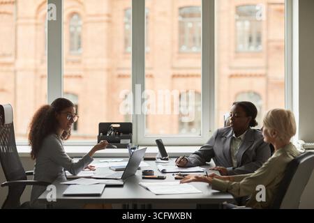 Three diverse businesswomen sitting at office table discussing documents and using laptop during professional meeting near large windows Stock Photo
