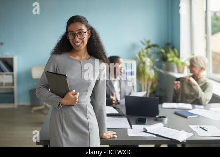 Portrait of young adult biracial woman smiling at camera while standing in modern office holding digital tablet, two diverse women working together at desk in background Stock Photo
