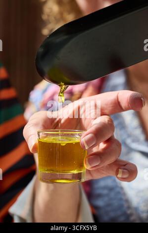 Close up of a woman's hand holding a small glass while pouring olive oil from a metal scoop, during an olive oil tasting Stock Photo