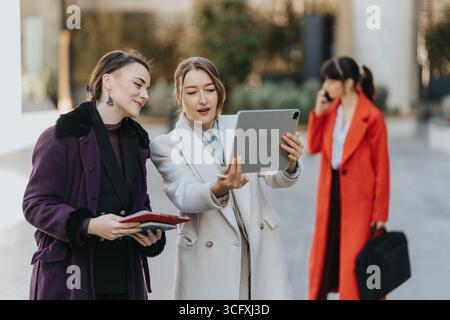 Two businesswomen collaborate outdoors using a digital tablet, interacting and discussing work together. Stock Photo