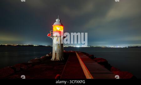 Radiating red light from a lighthouse, guiding ships through the dark night, while distant city lights blur softly on the horizon Stock Photo
