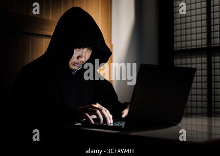 A hooded man works on a laptop in a dimly lit room, symbolizing hacker activity, cybercrime, data theft, and online security threats in the digital ag Stock Photo