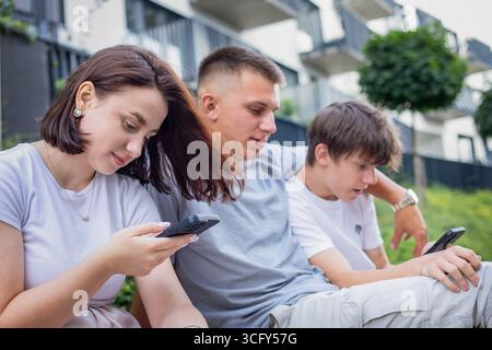 Three young people sitting on a bench in the park, using smartphones and browsing social media while relaxing outdoors. High quality photo Stock Photo