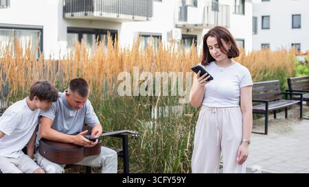 Three young people sitting on a bench in the park, using smartphones and browsing social media while relaxing outdoors. High quality photo Stock Photo