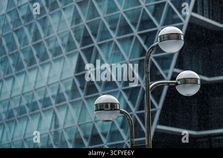 Close-up three white globe shaped streetlights in rain sitting on curved metal arms varying in height against backdrop of geometric patterned glass. Stock Photo