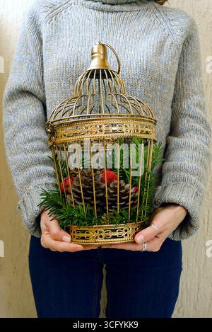 Christmas home decor: woman holding birdcage with pine cones and evergreen branches. DIY project using natural materials and upcycled vintage items Stock Photo