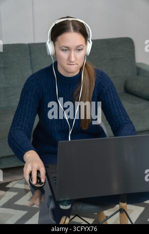 Young woman wearing headphones, engaging in a video call while working from her home office. Focused on her laptop, she talks and navigates with a com Stock Photo
