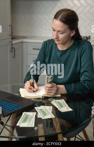 Focused woman writing notes in notebook, managing home budget, calculating money and making financial plan while sitting at kitchen table with laptop Stock Photo