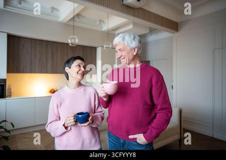 Happy elderly couple drinking tea together. Laughing and talking at home, joyful retirement moment. Stock Photo
