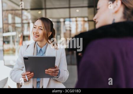 Two businesswomen collaborating outdoors, using a tablet device, and engaging in a professional yet approachable discussion in a modern urban setting. Stock Photo