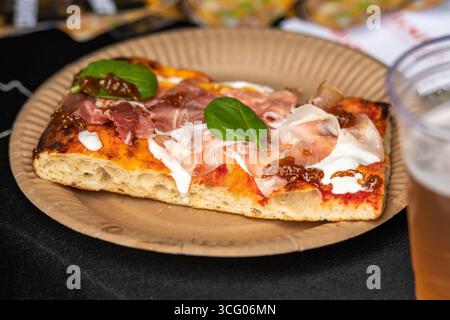 A slice of pizza topped with fresh basil leaves, prosciutto crudo and stracciatella. The pizza is served on a paper plate with a glass of beer Stock Photo