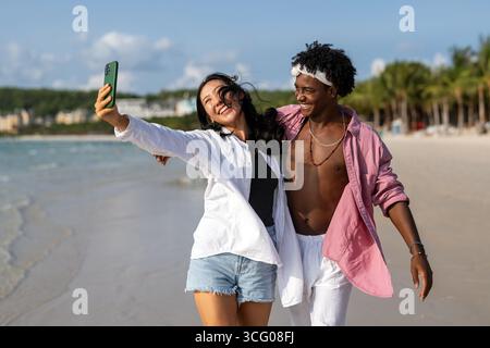 Trendy digital creator couple taking a stylish selfie by the beach, sharing travel content for social media, living the influencer lifestyle in paradi Stock Photo