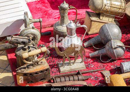 Collection of old household items, traditional metalware, antique tools, and decorative objects displayed on a red carpet at a flea market in Riyadh, Stock Photo