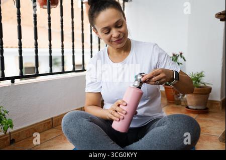A cheerful woman seated on a balcony holding a pink water bottle. The relaxed atmosphere and greenery accentuate the tranquil mood of the environment. Stock Photo