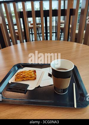 A paper cup filled with coffee sits next to a pastry on a tray, placed on a wooden table in a contemporary cafe atmosphere with seating Stock Photo