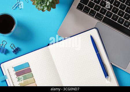 Laptop, notebook, pen, coffee, and plant on blue desk. Minimal workspace with space for your text. Stock Photo