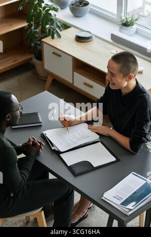 Vertical high angle view shot of cheerful young Caucasian woman sitting at desk in office having conversation with African American job applicant Stock Photo