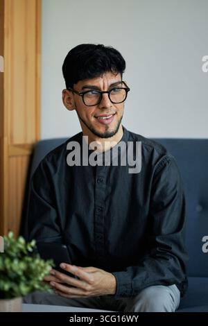 Vertical medium portrait of handsome young Hispanic man wearing eyeglasses sitting on couch in modern office holding smartphone looking away Stock Photo