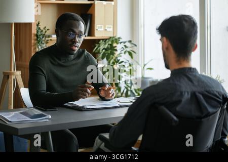 Young African American manager and his Hispanic co-worker with disability sitting at desk in front of each other working together on business strategy Stock Photo