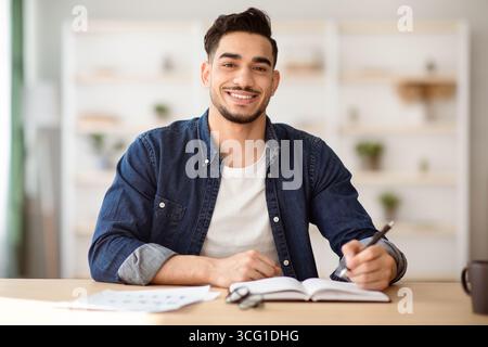 Cheerful arab guy in casual sitting at workdesk, taking notes Stock Photo