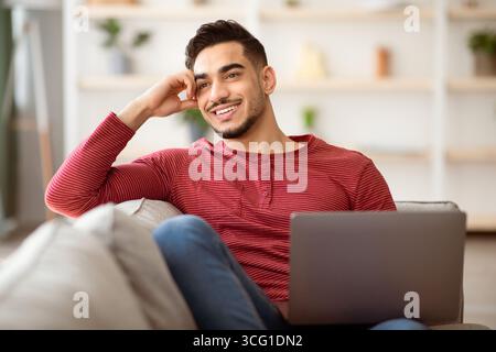 Dreamy arab guy resting on couch at home, using laptop Stock Photo