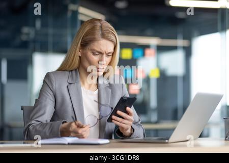 A professional woman in business attire is focused on her smartphone in a modern office setting. She appears puzzled or concerned while reviewing info Stock Photo