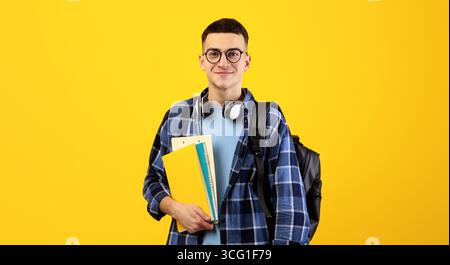Education concept. Positive young man with backpack, headphones and notebooks posing on orange background Stock Photo