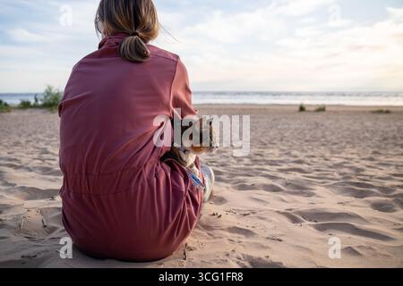 Beach holiday with pet. Woman relaxing on sandy shore with leashed cat during morning walk rearview Stock Photo