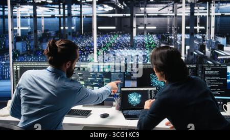Data center admins doing brainstorming, working on neural network software development. Server farm IT employees working together, implementing machine learning technology using PC, camera A Stock Photo