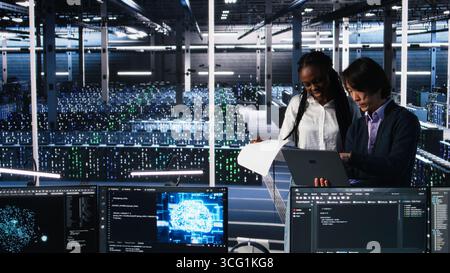 Data center engineers managing AI systems for uptime and efficiency, looking over paperwork files. African american woman and Asian man reading documents in multiethnic server hub, camera A Stock Photo