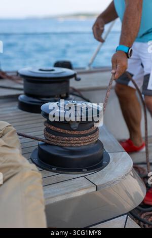 A man on a sailboat pulls the sheet to adjust the sail. A close-up of a winch and hands at work, representing the action and passion of sailing. Stock Photo